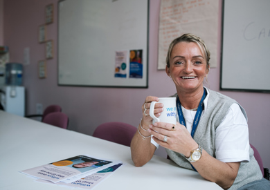 A smiling woman with a lanyard sits at a table holding a coffee mug, with informational pamphlets and a whiteboard visible in the background.