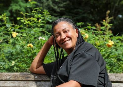 A joyful woman with braided hair sits on a park bench, smiling at the camera, with sunlit trees and yellow flowers in the background.