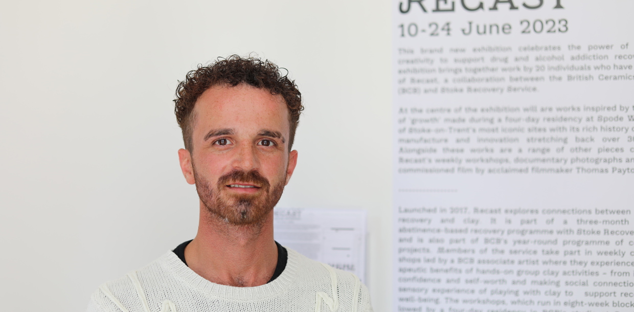 A man with curly hair and a beard smiles at the camera, standing in front of a white exhibition wall.