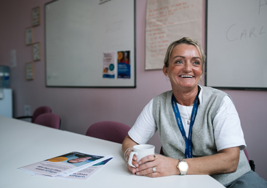 A smiling woman with a lanyard sits at a table holding a coffee mug, with informational pamphlets and a whiteboard visible in the background.
