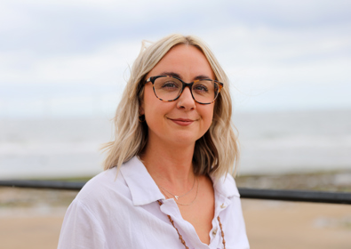 A smiling woman with shoulder-length blonde hair and round glasses, wearing a white shirt, stands outdoors with a beach background.