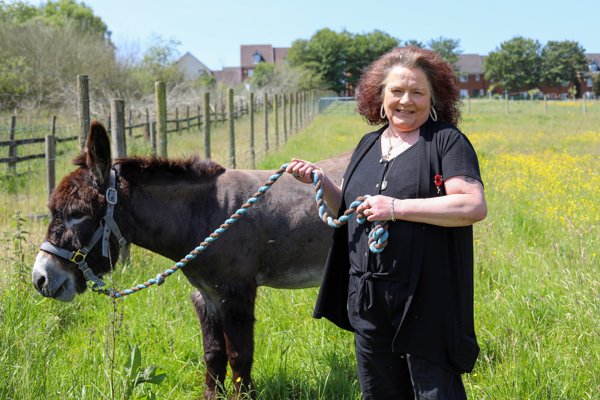 A woman in a black shirt stands smiling in a field, holding a leash attached to a calm donkey.