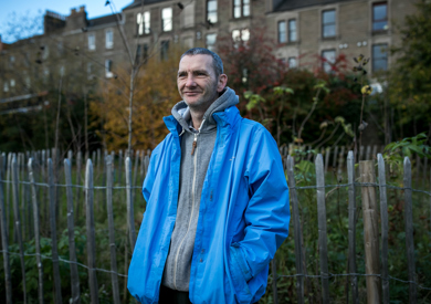 A man in a blue jacket and gray hoodie smiles gently, standing outdoors with an old building and trees in the background.