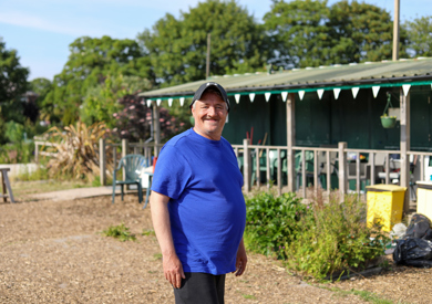 A cheerful middle-aged man in a blue t-shirt standing in a community garden with greenery and a small shed in the background.