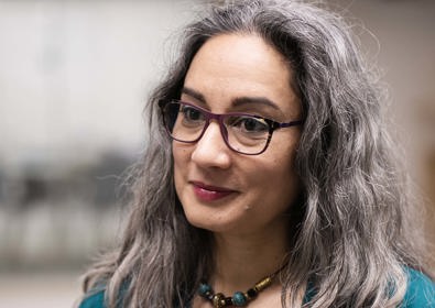 A woman with shoulder-length gray hair and glasses, wearing a teal top and black cardigan, in an office setting.