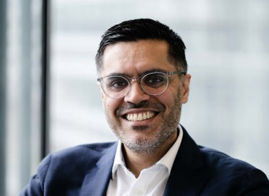 Portrait of a smiling man wearing a navy blazer and white shirt, sitting indoors with a window background that filters soft natural light.