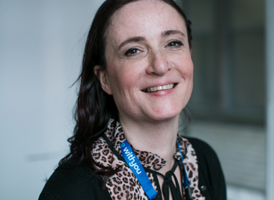 A smiling woman with dark hair tied back, wearing a leopard print blouse and a lanyard, standing in an office environment.