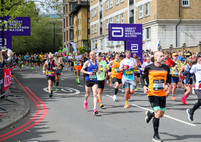 Runner wearing a WithYou vest participating in a city marathon, with spectators along the barricaded course. signs and banners are visible.