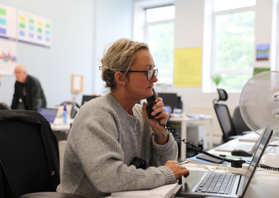 A woman in a gray sweater speaks on a telephone in a bright, busy office environment, with computers and a colleague visible in the background.