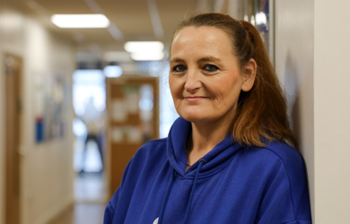 A middle-aged woman, wearing a blue hoodie, stands near a hallway wall, smiling gently towards the camera.