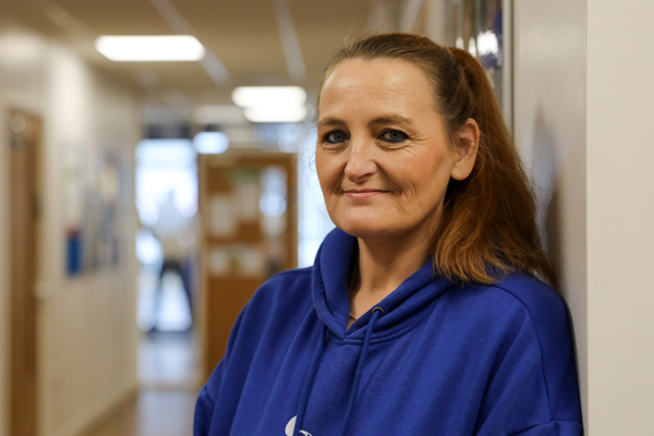 A middle-aged woman, wearing a blue hoodie, stands near a hallway wall, smiling gently towards the camera.