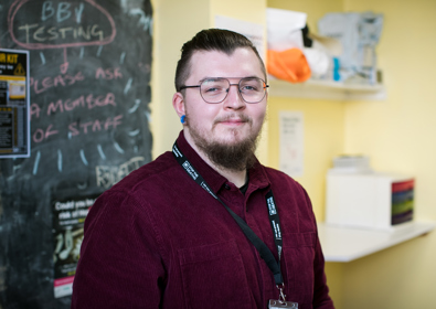 A man with glasses and beard, dressed in a burgundy shirt, stands smiling in a room adorned with health-related posters and notices. 