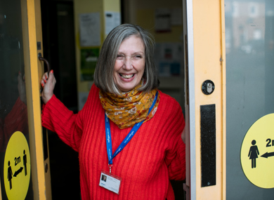 A cheerful woman with gray hair, wearing a red sweater and colorful scarf, stands at an open door smiling, with a lanyard and id around her neck.