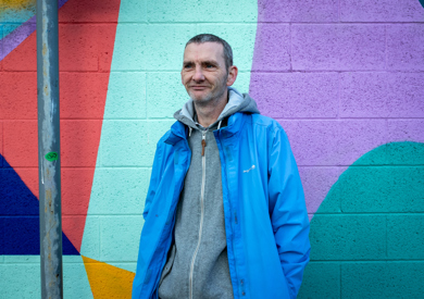 A man in a blue jacket standing in front of a colorful, geometric mural.