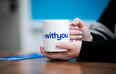 Close-up of a person's hands holding a white mug with the words "Withyou" in blue text.