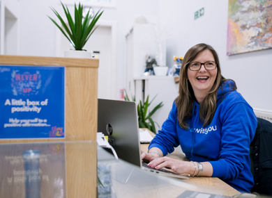 A woman in a blue hoodie smiling at a reception desk with a laptop, motivational signs, and a plant in a modern office setting.