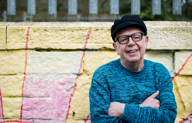 An smiling elderly man in glasses and a black cap. He sits before a colourfully painted wall.