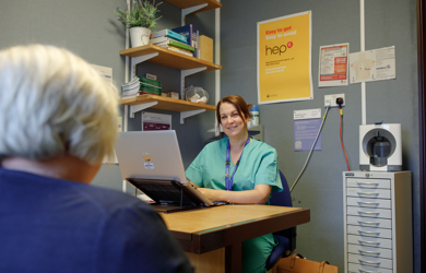 A female nurse in scrubs sits at a desk with a laptop, smiling at an elderly patient whose back is to the camera, in a clinic office decorated with informational posters.