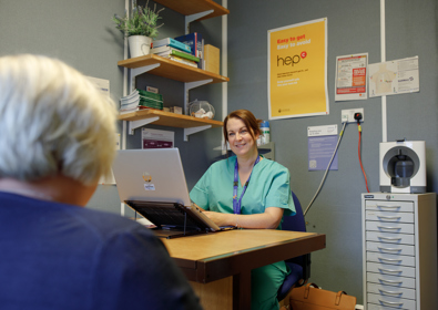A female nurse in scrubs sits at a desk with a laptop, smiling at an elderly patient whose back is to the camera, in a clinic office decorated with informational posters.