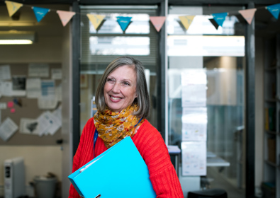 A smiling woman with grey hair, wearing a red sweater and a colourful scarf, holds a light blue folder in a room with glass doors in the background.
