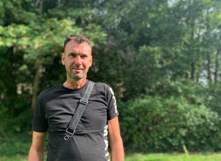 A man standing outdoors with trees in the background. He wears a black and is smiling.