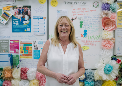 A smiling woman in a white blouse stands in front of a colorful community bulletin board filled with flyers, drawings, and decorative flowers.