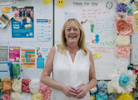 A smiling woman in a white blouse stands in front of a colorful community bulletin board filled with flyers, drawings, and decorative flowers.