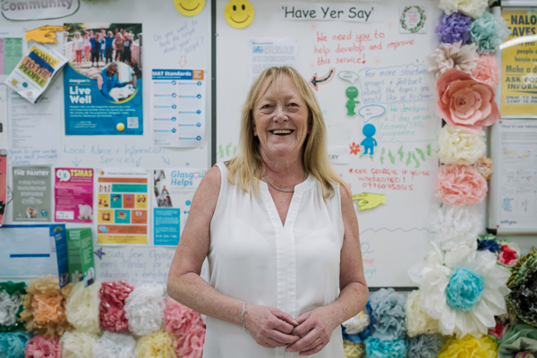 A smiling woman in a white blouse stands in front of a colorful community bulletin board filled with flyers, drawings, and decorative flowers.