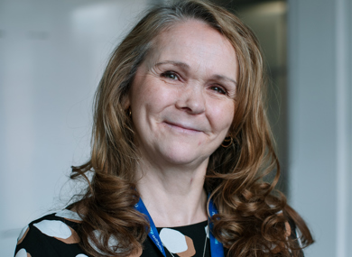 A portrait of a smiling woman with long, curly brown hair, wearing a black and white polka-dot blouse and a blue lanyard, standing indoors.