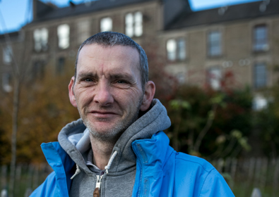 A man in a blue jacket and gray hoodie smiles gently, standing outdoors with an old building and trees in the background.