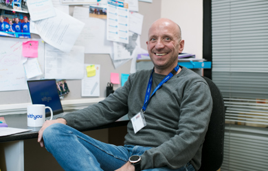 A smiling man sits at a desk in an office filled with papers and photographs, wearing a grey sweater and a blue lanyard.