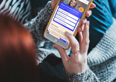 Person in gray sweater holding a phone displaying the WithYou support website, sitting on a zebra-patterned blanket.
