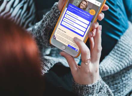 Person in gray sweater holding a phone displaying the WithYou support website, sitting on a zebra-patterned blanket.