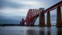 Shot of bridge, a cantilever railway bridge over a body of water under cloudy skies, highlighting its red colour and massive stone piers.