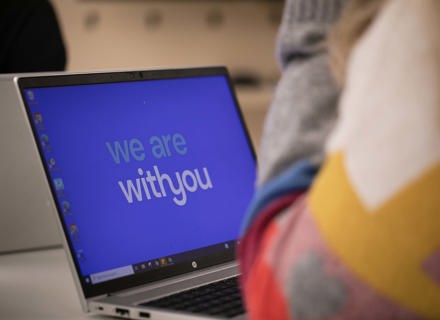 A laptop screen displaying the message "we are with you" in white text on a blue background, partially obscured by a person in a pastel-coloured sweater sitting in the foreground.