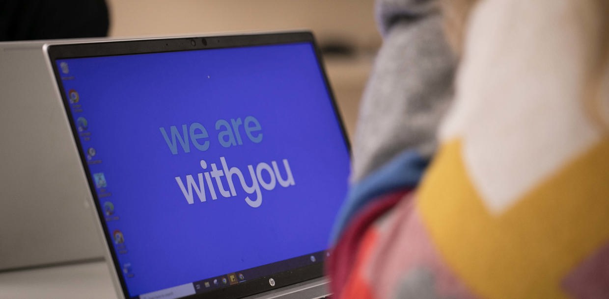 A laptop screen displaying the message "we are with you" in white text on a blue background, partially obscured by a person in a pastel-coloured sweater sitting in the foreground.