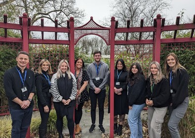 A group comprising of five women and four men, standing in front of a decorative red garden gate, all smiling at the camera.