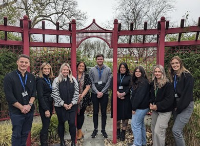 A group comprising of five women and four men, standing in front of a decorative red garden gate, all smiling at the camera.