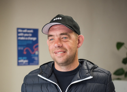 A smiling man wearing a black jacket and cap stands indoors, with a blue WithYou notice board visible in the background.