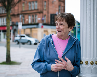 An older woman in a blue hoodie and pink shirt smiles and gestures with her hands while standing next to a white street lamp post in a city setting.