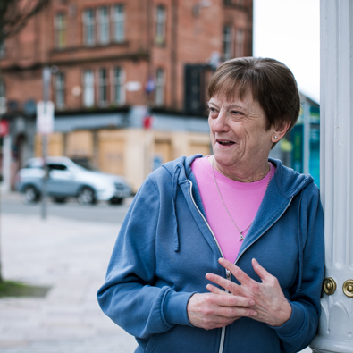 An older woman in a blue hoodie and pink shirt smiles and gestures with her hands while standing next to a white street lamp post in a city setting.