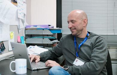 A smiling man sits at a desk in an office filled with papers and photographs, wearing a grey sweater and a blue lanyard.