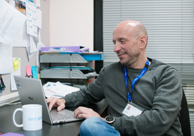 A smiling man sits at a desk in an office filled with papers and photographs, wearing a grey sweater and a blue lanyard.