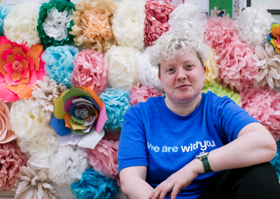 A woman in a blue t-shirt sits smiling in front of a colorful background of assorted paper flowers in various sizes and colors.