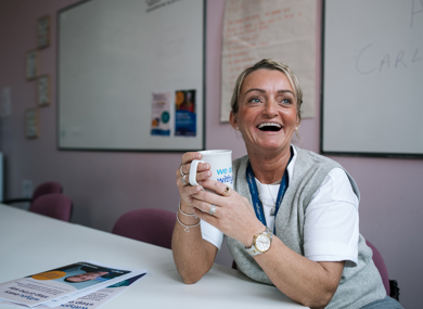 A smiling woman with a lanyard sits at a table holding a coffee mug, with informational pamphlets and a whiteboard visible in the background.