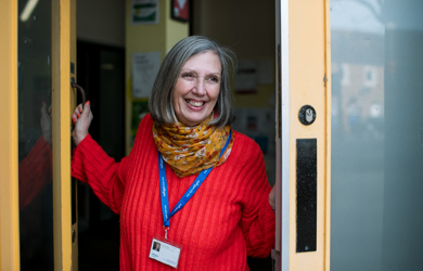 A cheerful woman with gray hair, wearing a red sweater and colorful scarf, stands at an open door smiling, with a lanyard and id around her neck.