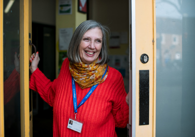 A cheerful woman with gray hair, wearing a red sweater and colorful scarf, stands at an open door smiling, with a lanyard and id around her neck.