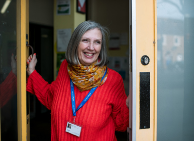 A cheerful woman with gray hair, wearing a red sweater and colorful scarf, stands at an open door smiling, with a lanyard and id around her neck.