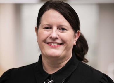 Portrait of a smiling woman wearing a black shirt and a metallic necklace, standing in an office setting with a softly blurred background.