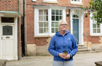 A middle-aged woman wearing glasses and a blue hoodie stands in front of a brick building holding a coffee cup. she appears thoughtful and content.